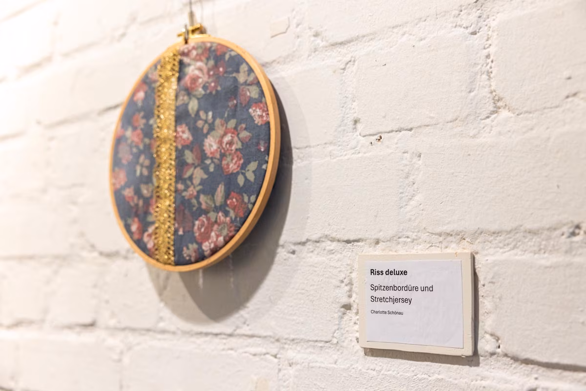 An der Wand hängt ein mit Stoff bespannter Rahmen, wo ein Riss mit Spitzenbordüre abgedeckt ist. Daneben ein Museumsschild, welches die Flicktechnik beschreibt.