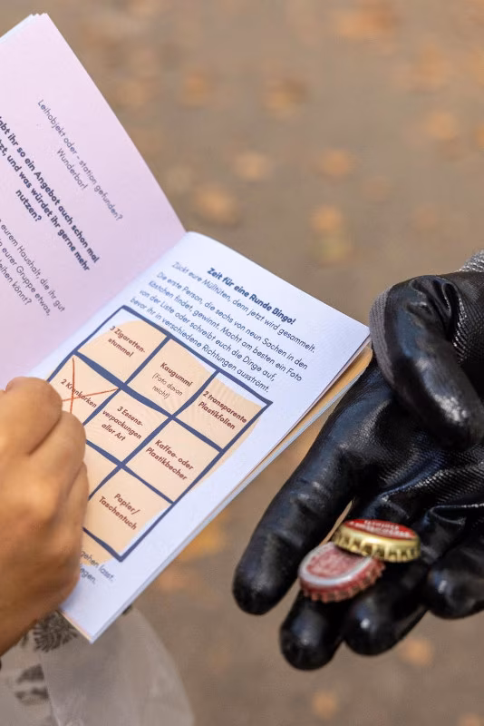 Ein Blick ins Schweif-Heft, das Müll-Bingo ist zu sehen. Danaben hält eine Hand mit Handschuh ein paar Kronkorken ins Bild.