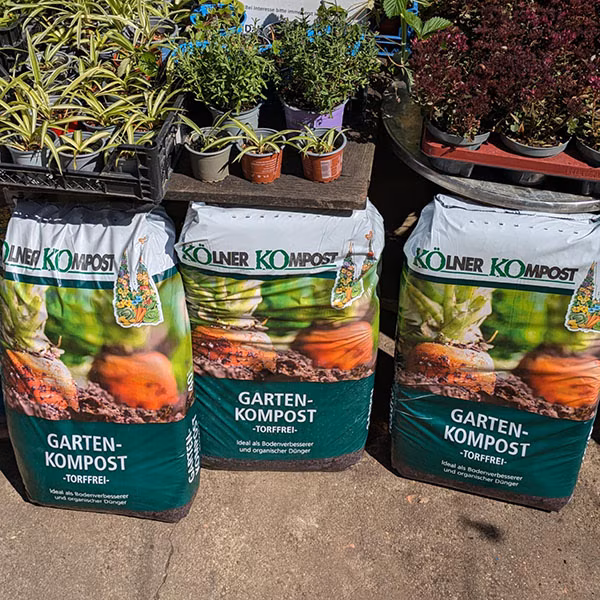 Drei Säcke mit Kölner Kompost, darauf stehen Pflanzen zum Verkauf in Töpfen
