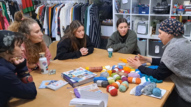 Jana erklärt vier Workshopteilnehmerinnen die Grundlagen vom Häkeln. Alle sitzen um einen Tisch voller Wolle, im Hintergrund sieht man die Einrichtung vom IGLU.