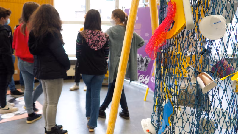 Im Vordergrund ist der Zero Waste Parcours zu erkennen - einige Plastikteile hängen in einem Netz. Im Hintergrund schaut sich eine Gruppe Schüler:innen eine Stellwand an und diskutiert.