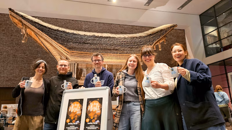 Team Zero Waste Köln hält Tassen in der Hand, die sie beim Quiz der Fair Trade Night gewonnen haben. Im Hintergrund der historische indonesische Reisspeicher im Foyer des Rautenstrauch-Joest-Museum am Neumarkt