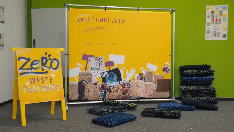 Links ein gleuchtend gelbes Schild "Zero Waste Parcours". In der Mitte eine Stellwand "Eure Stimme zählt". Drumherum sind Kissen ausgestapelt, die für eine Diskussion genutzt werden können.