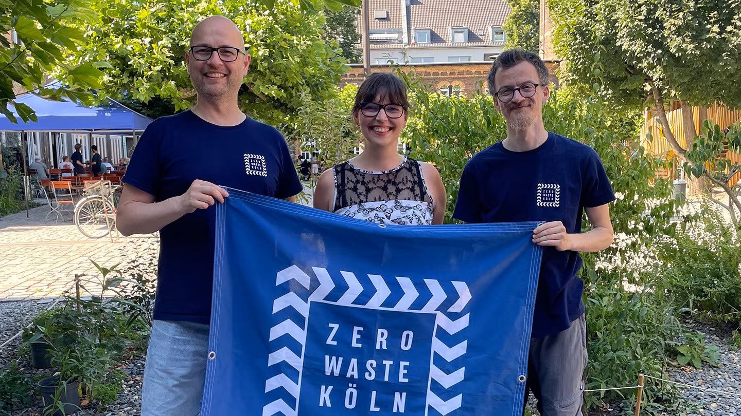Der neue Vorstand von Zero Waste Köln Franz, Roling, Pia Wroblewski und Jan Odenthal stehen im Innenhof der Alten Feuerwache und halten das Zero Waste Köln Banner in die Kamera