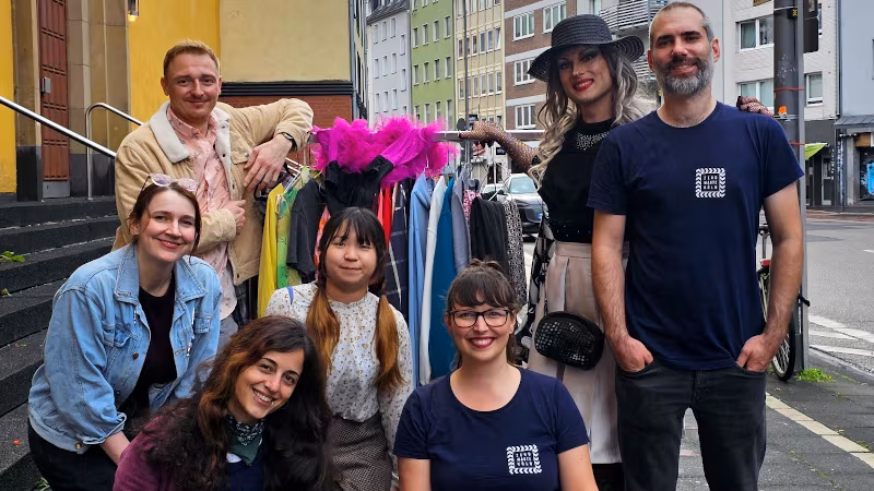 Das Team von Zero Waste Köln steht mit T-Shirts mit Logo auf der Straße. Etwas im Hintergrund ein Kleiderständer. Zu sehen ist auch die Dragtivistin Tricia Trash.