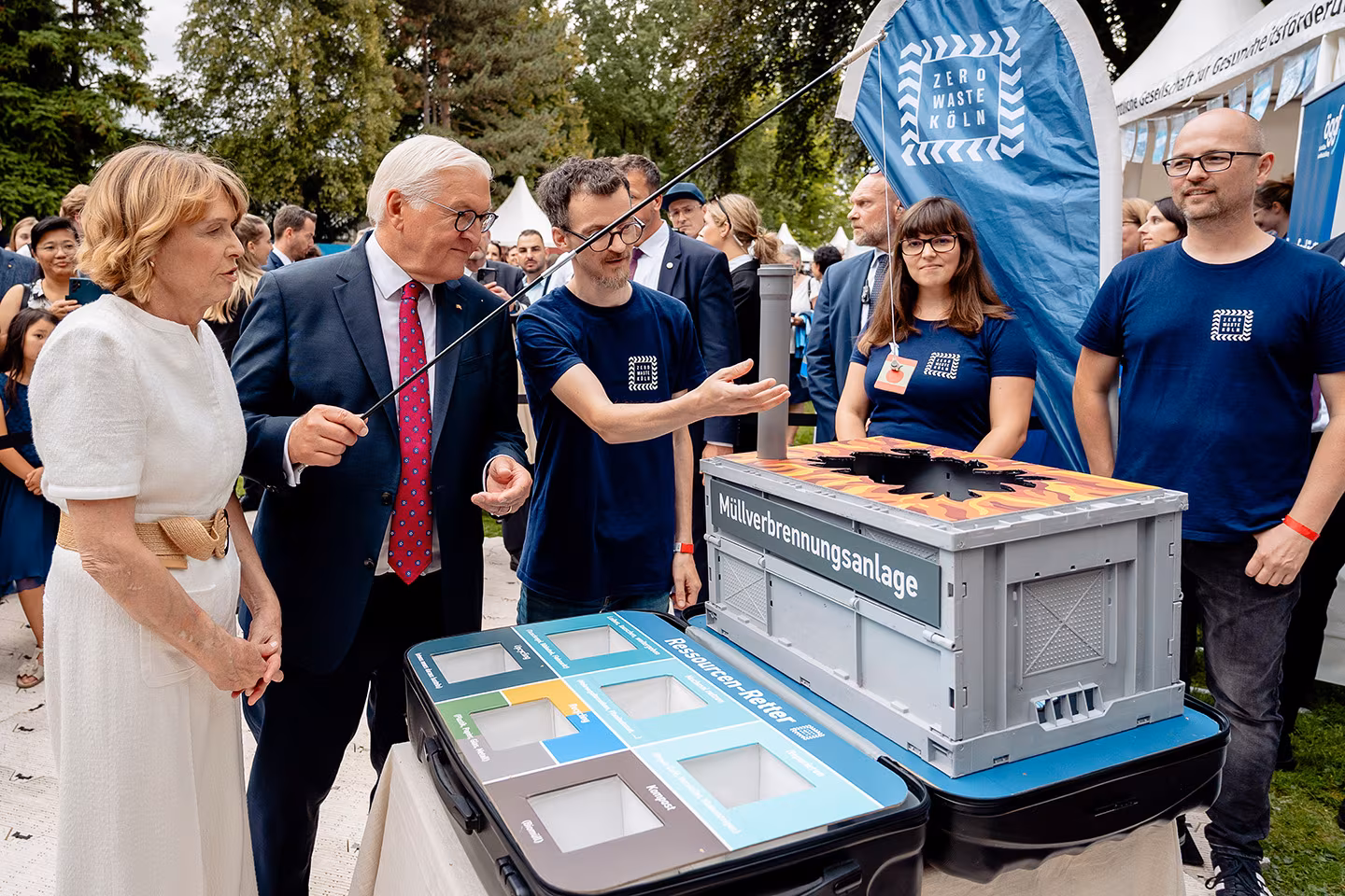 Der Bundespräsident steht mit seiner Ehefrau am Stand von Zero Waste Köln. In seiner Hand hält er eine magnetische Angel mit der er verschiedene Müllstücke aus einer selbstgebastelten Müllverbrennungsanlage angelt.